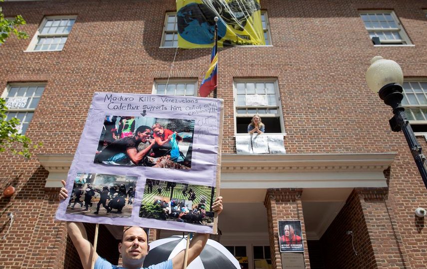 Desde la ventana una de las personas que ocupa la embajada de Venezuela en Washington, mientras un joven sostiene una pancartas denunciando la dictadura de Maduro.&nbsp;